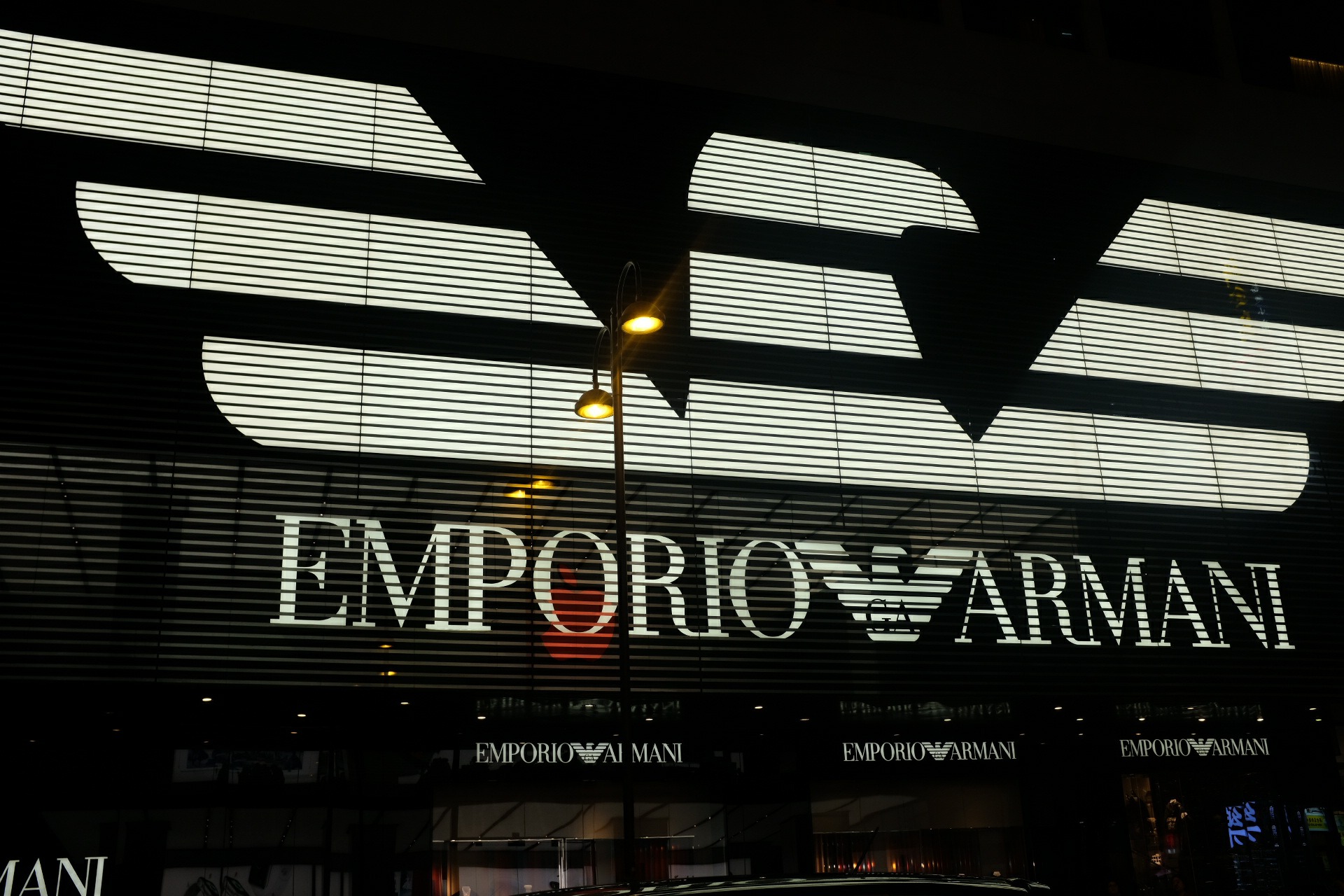 Illuminated Emporio Armani logo on a modern facade at night with geometric slats and a glowing streetlamp.