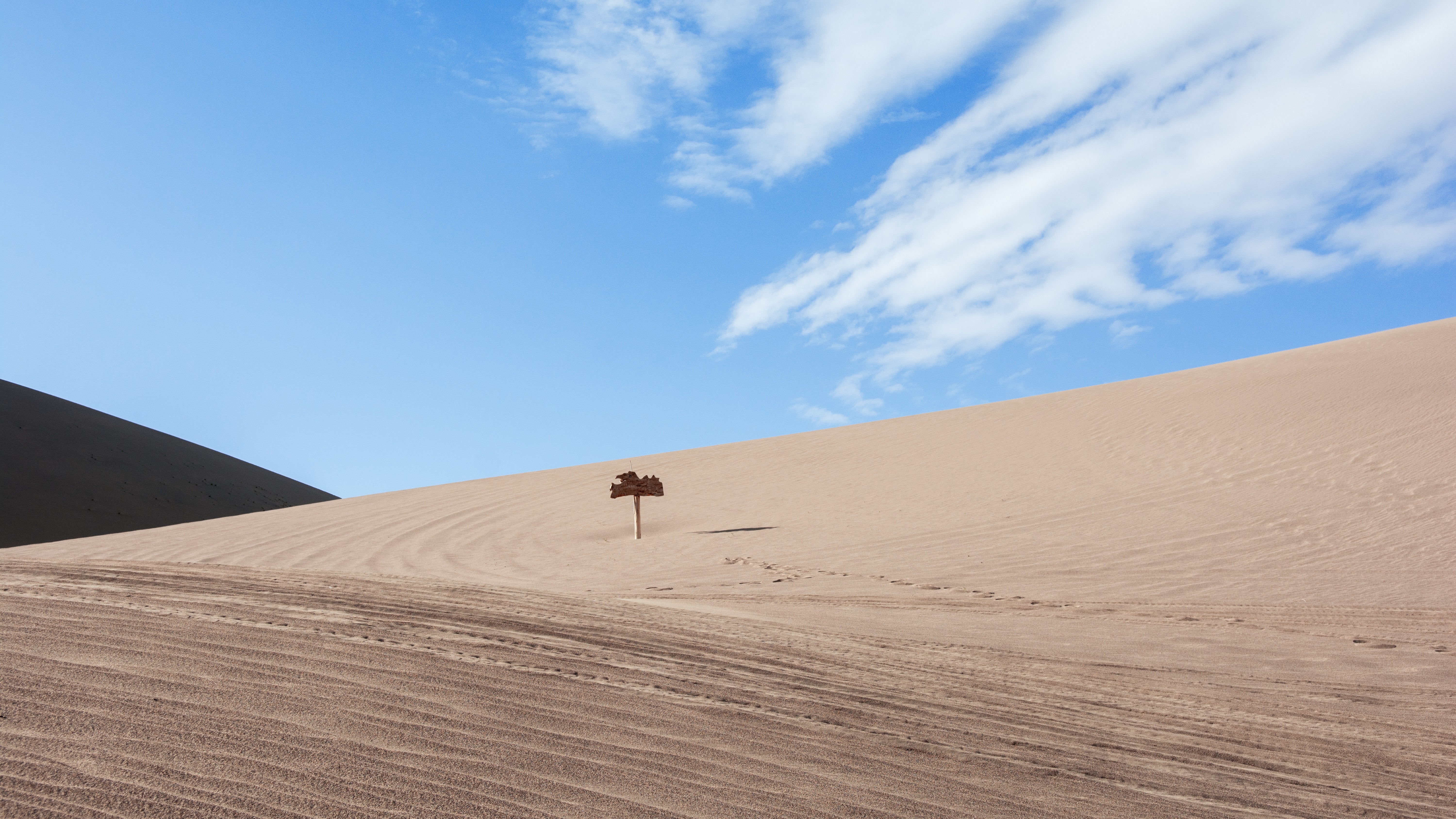 Weathered wooden signpost on a sunlit sand dune slope, featuring rippled desert terrain and a clear blue sky.