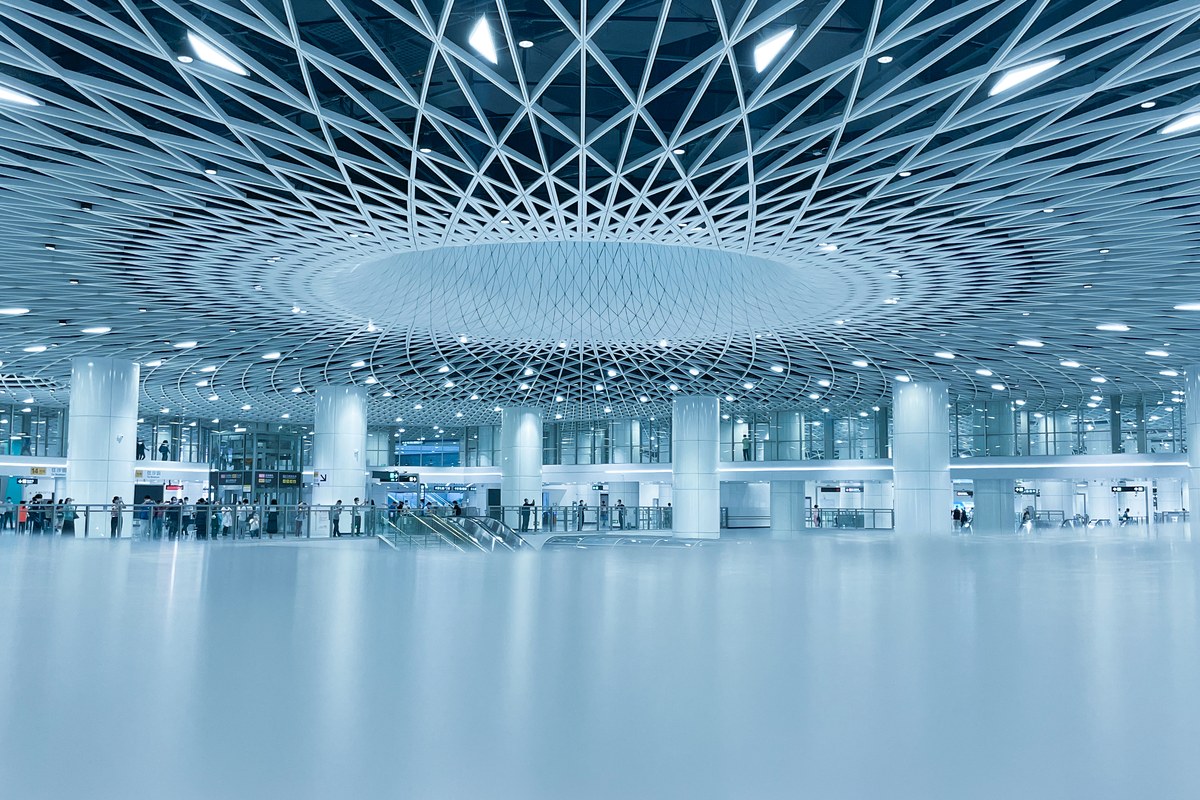 Modern airport terminal interior with a white geometric lattice ceiling, reflective floor, and travelers.