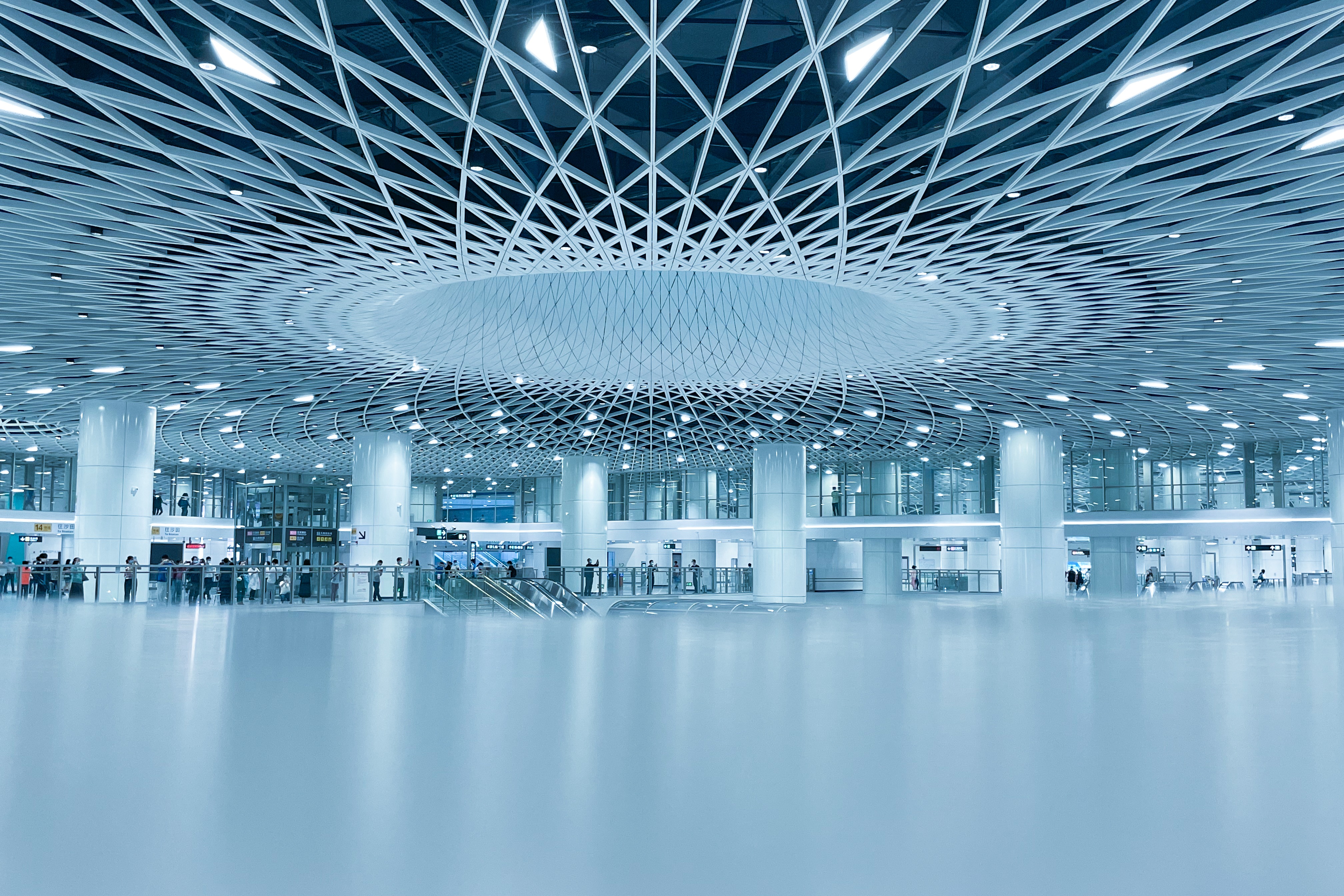 Modern airport terminal interior with a white geometric lattice ceiling, reflective floor, and travelers.