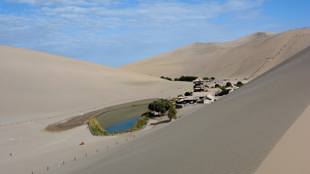 Wide-angle view of Yueyaquan crescent lake amid Mingsha Shan dunes, with pavilions and visitors under a clear sky.