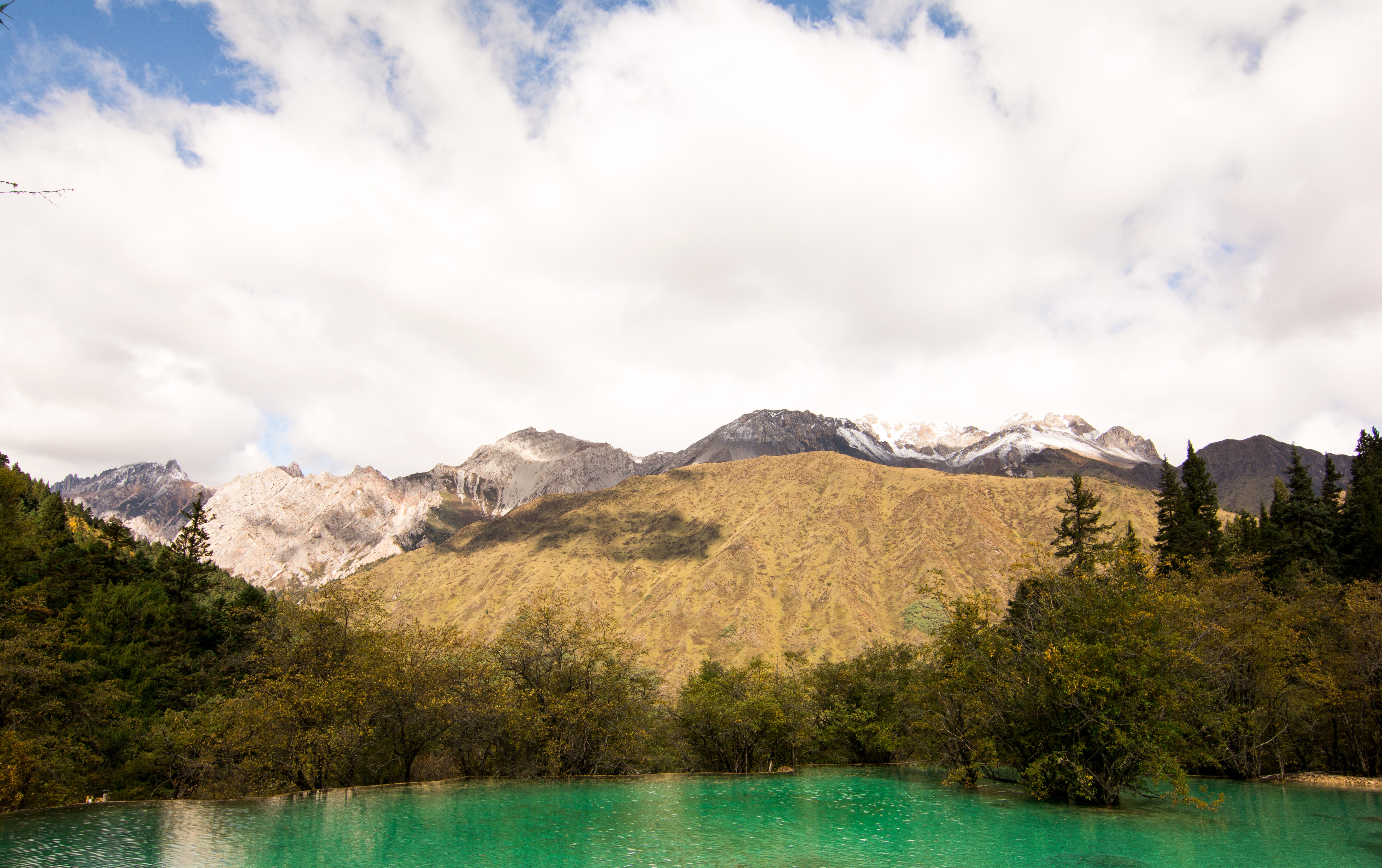 Wide-angle view of a turquoise alpine lake reflecting snow-capped mountains and golden autumn trees.