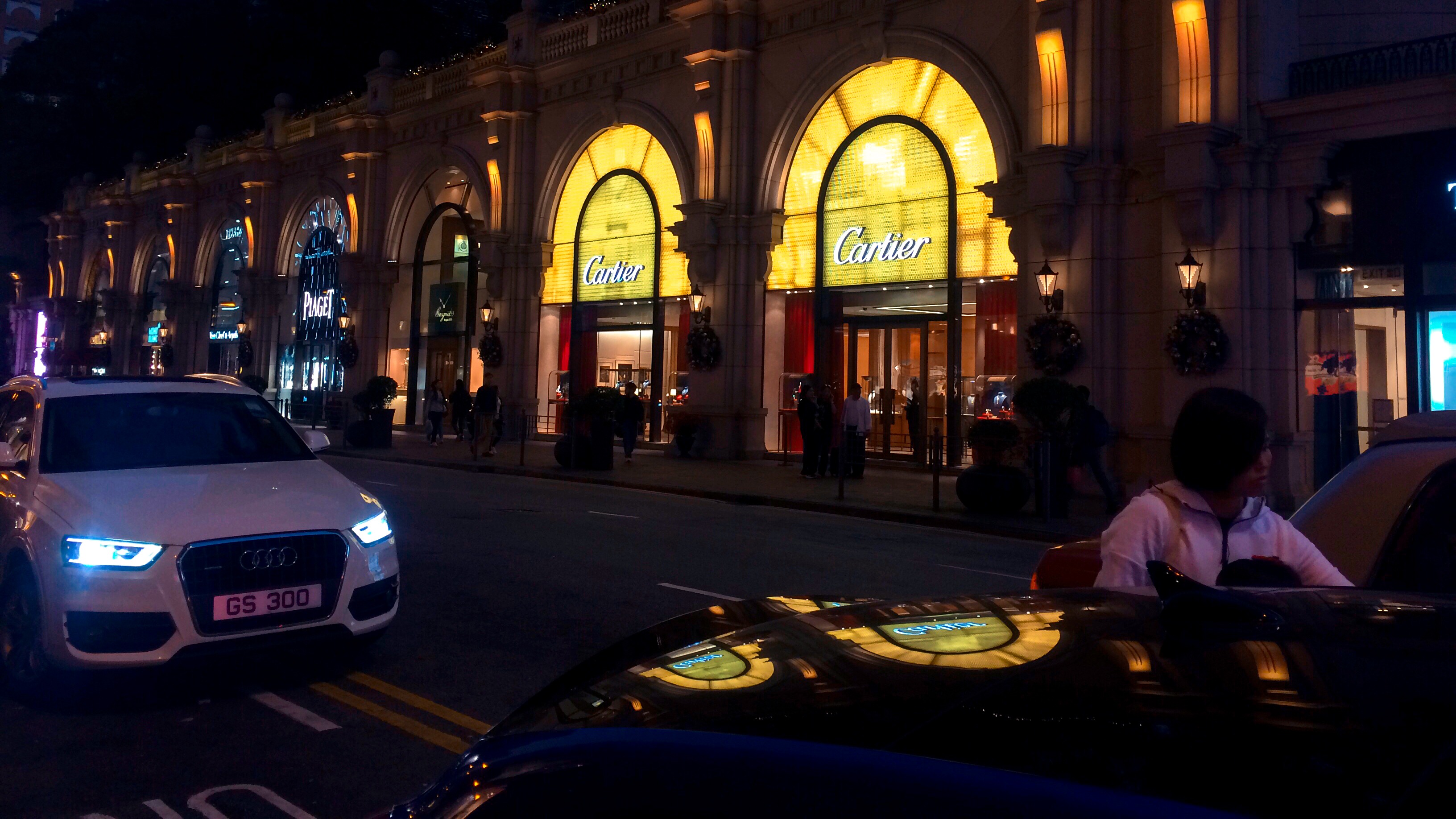 Low-angle night view of illuminated luxury retail facades in Hong Kong, with vehicles on a wet city street.