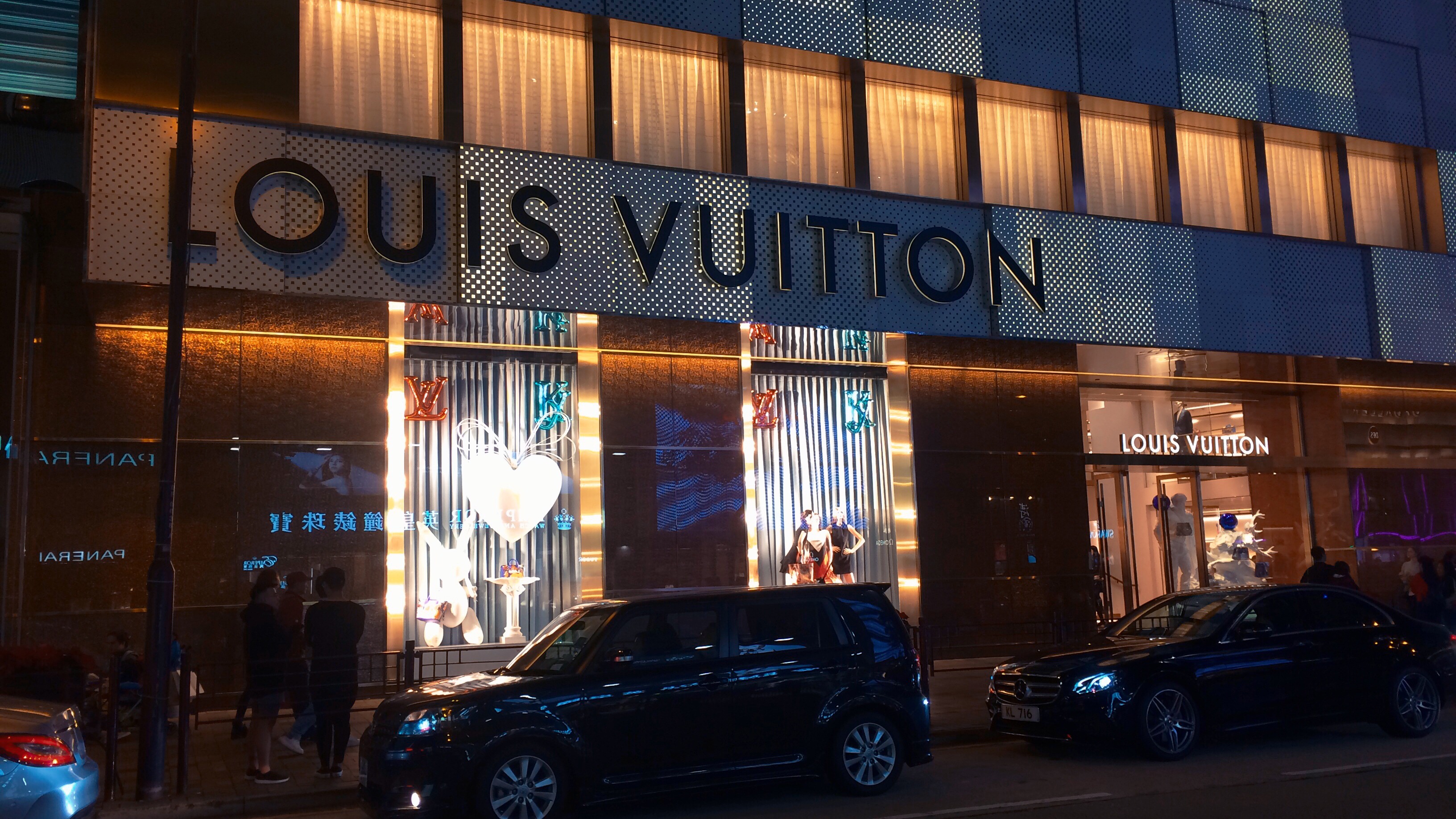 Nighttime view of the illuminated Louis Vuitton store facade in Hong Kong, featuring glowing windows and parked cars.