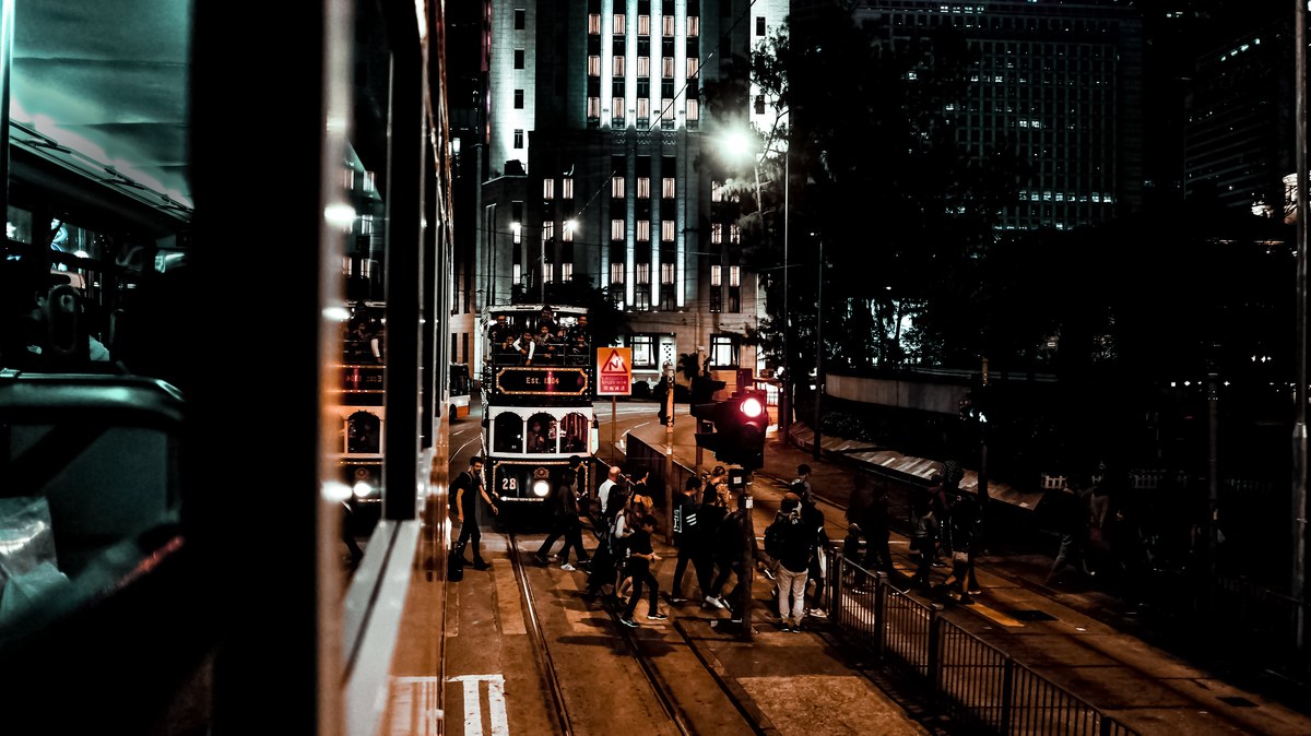 Vintage double-decker tram glowing on a Hong Kong street at night, framed by towering buildings and glass reflections.