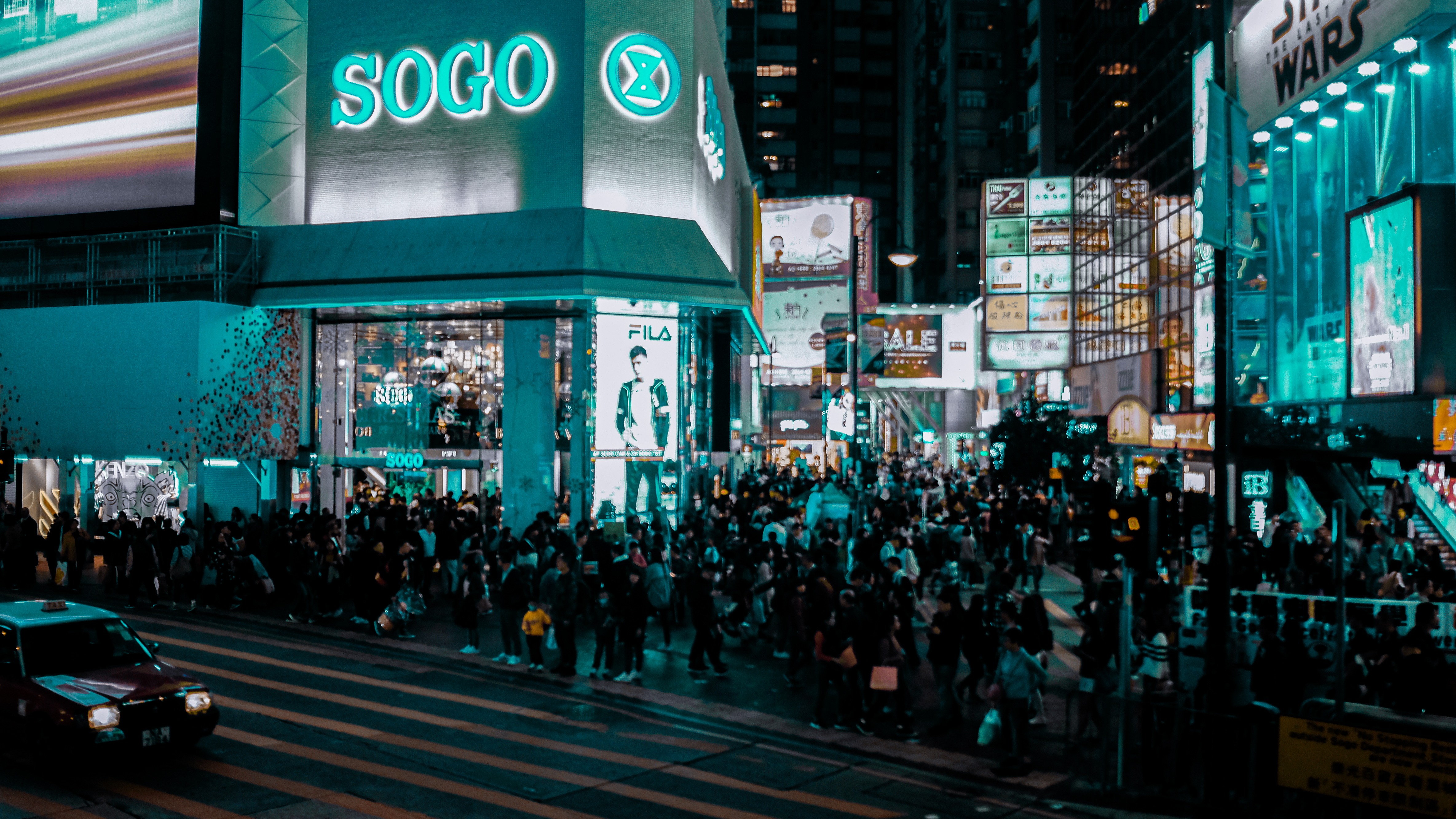Night street photo of SOGO Causeway Bay showing glowing teal neon signs, blurred pedestrians crossing, and a red taxi.