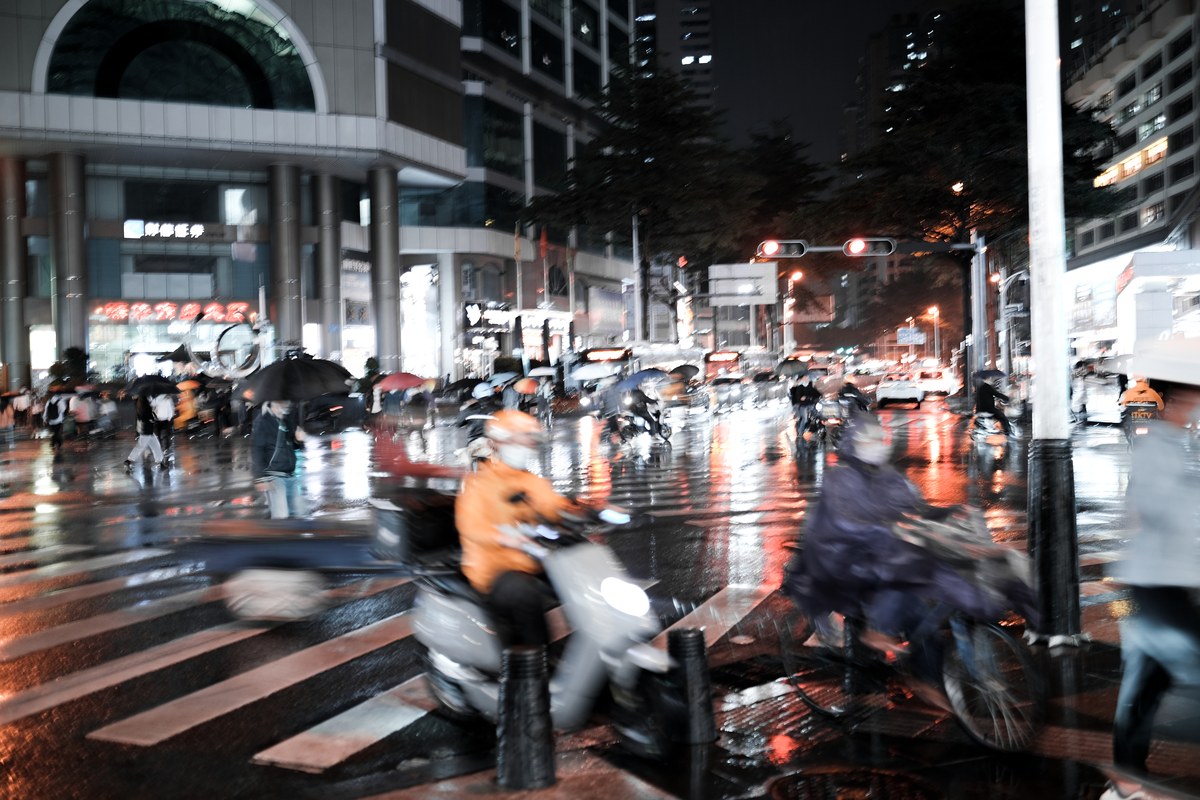 Rain-soaked Shenzhen crosswalk at night with blurred scooters, pedestrians, and glowing neon reflections on wet pavement.