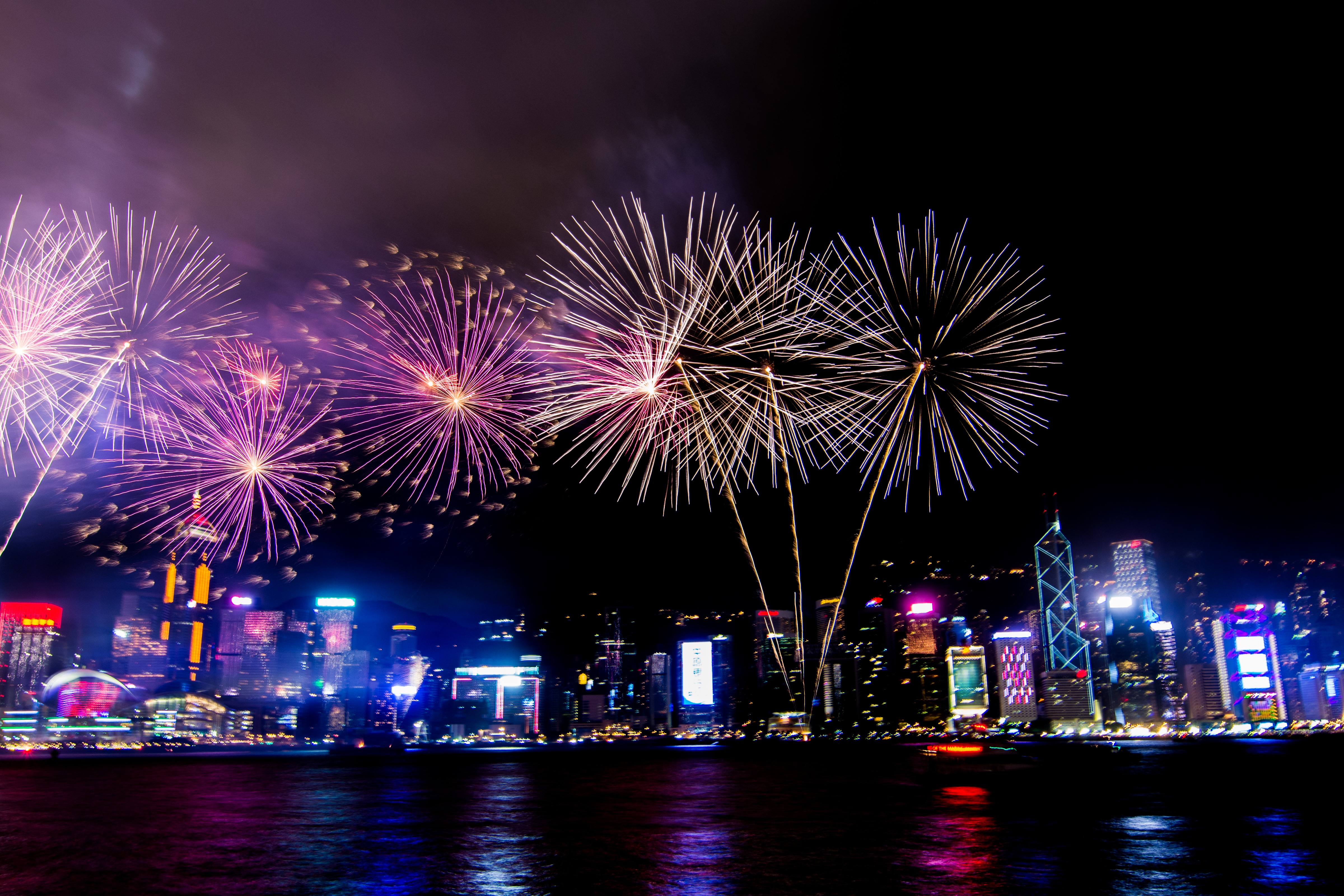 Colorful fireworks burst over a lit city skyline at night, reflecting on the calm harbor waters below.