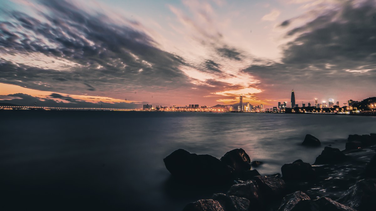 Panoramic twilight view of Shenzhen skyline reflected in calm coastal waters with silhouetted rocks and illuminated skyscrapers.