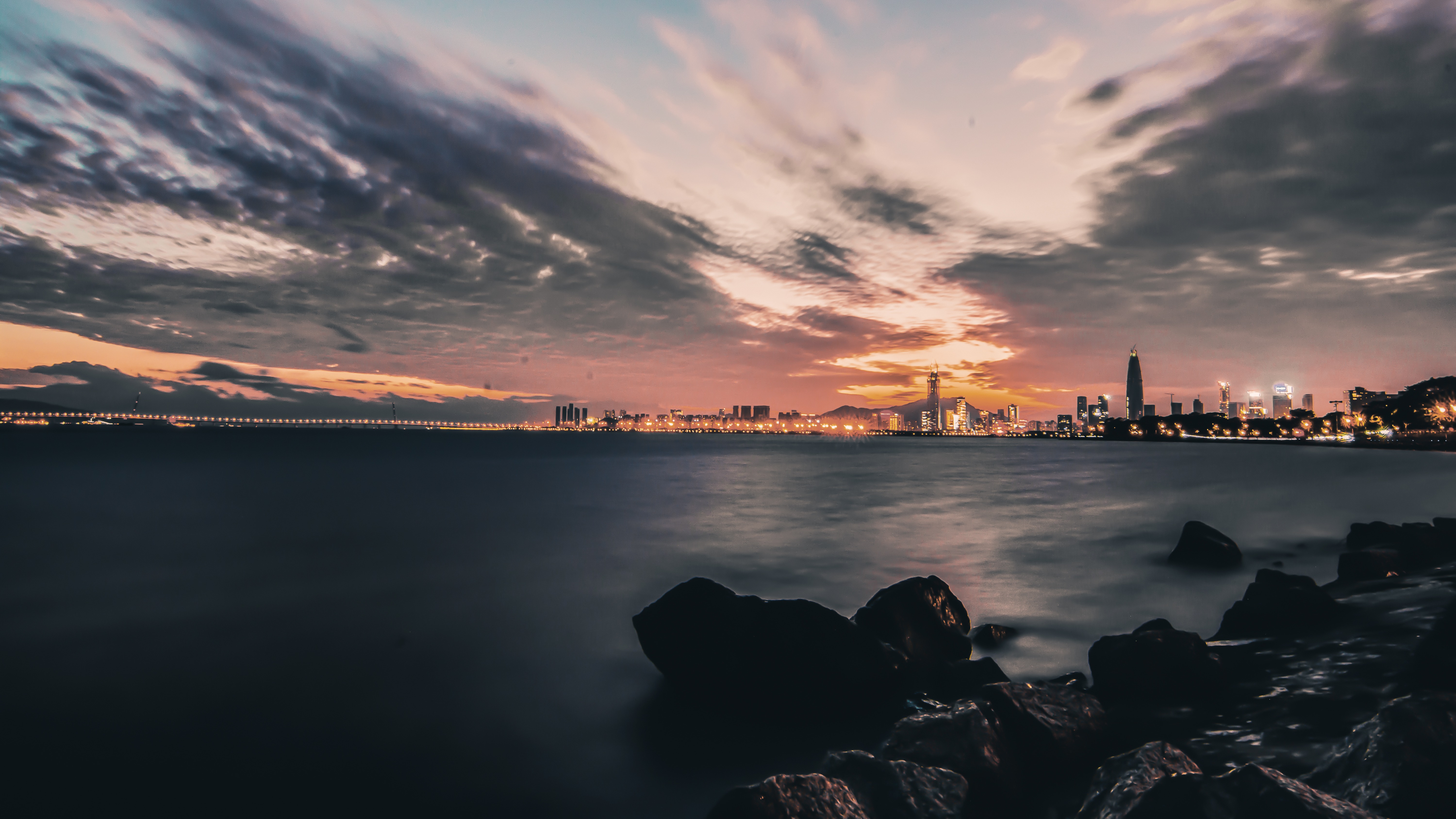 Panoramic twilight view of Shenzhen skyline reflected in calm coastal waters with silhouetted rocks and illuminated skyscrapers.