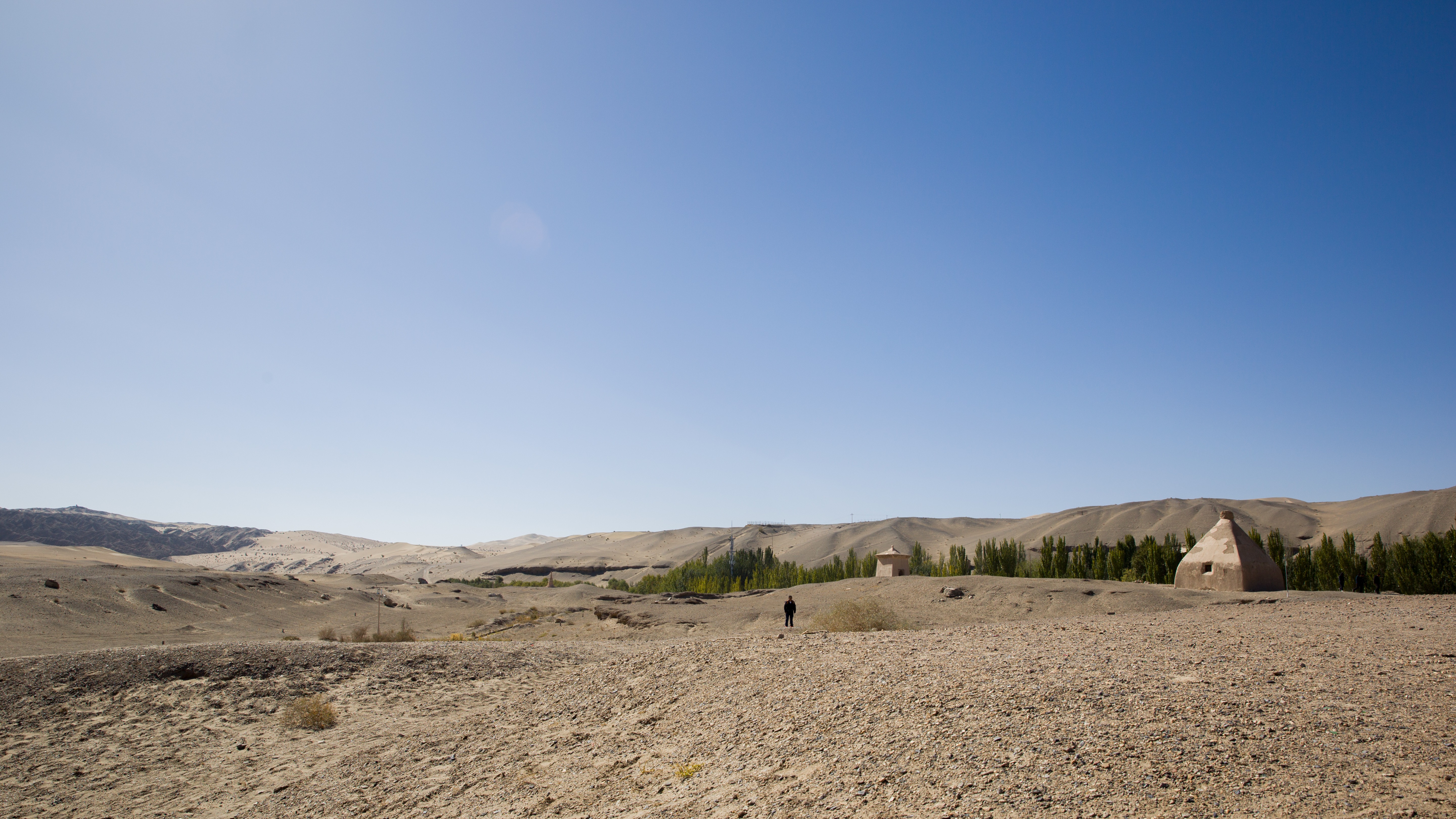 A solitary figure stands in a vast desert near a conical mud-brick tower under a clear blue sky.