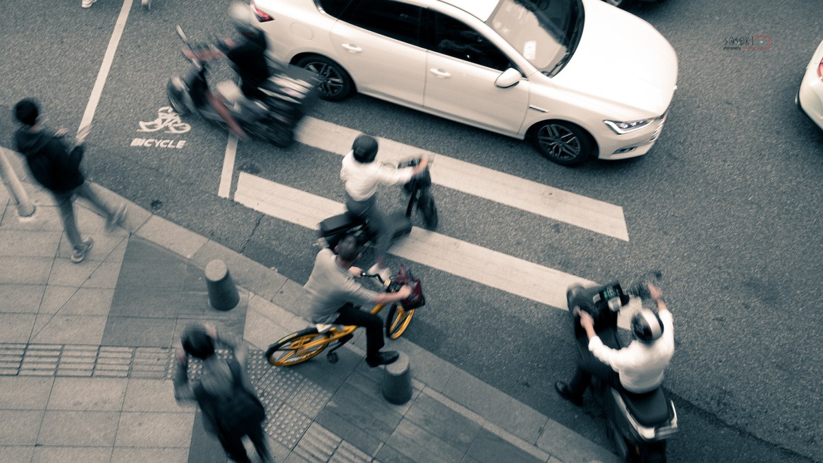 Bird's-eye view of an urban crosswalk with a yellow shared bike, blurred riders, and pedestrians on a muted street.