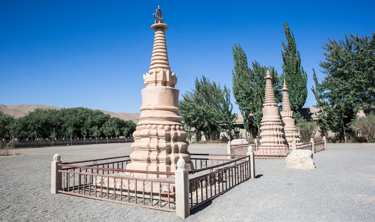 A tiered Buddhist stupa in a gravel courtyard under a clear blue sky, surrounded by smaller monuments and trees.