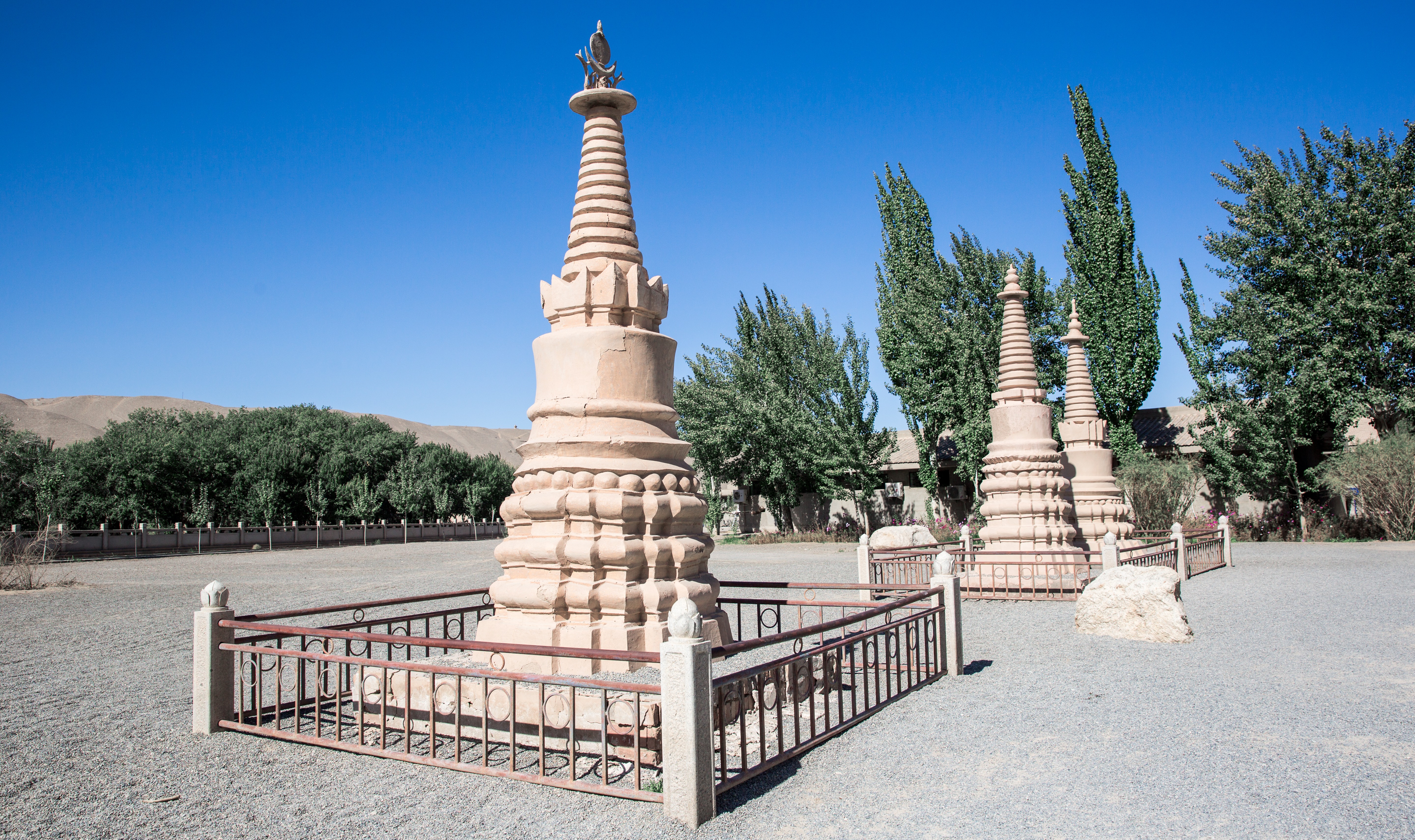 A tiered Buddhist stupa in a gravel courtyard under a clear blue sky, surrounded by smaller monuments and trees.