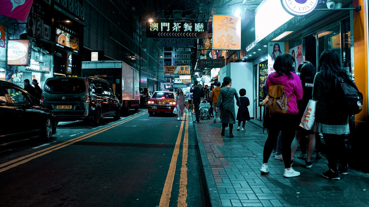 Pedestrians walking down a narrow Hong Kong alley at night, framed by glowing neon signs and parked cars on wet asphalt.