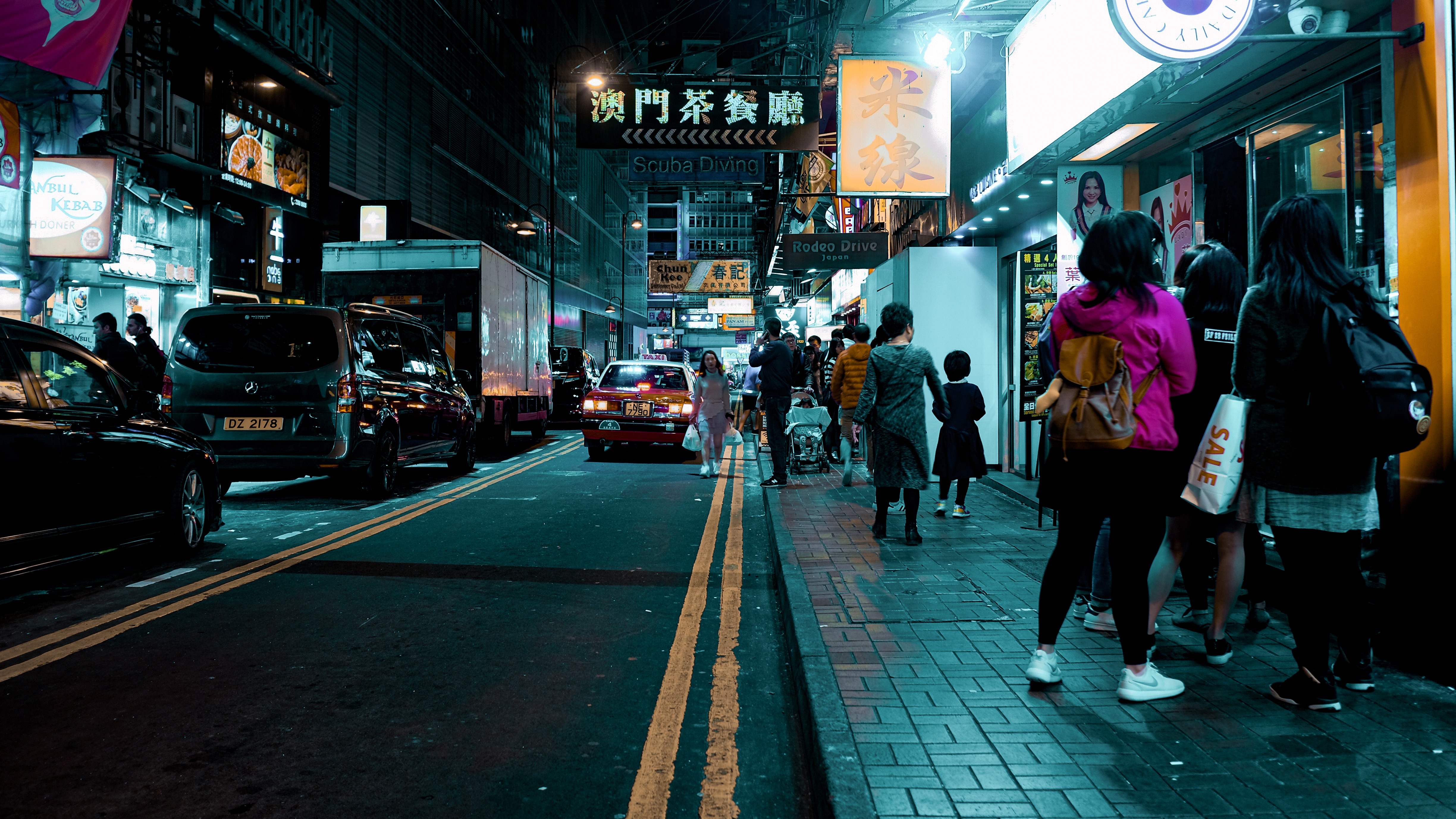 Pedestrians walking down a narrow Hong Kong alley at night, framed by glowing neon signs and parked cars on wet asphalt.